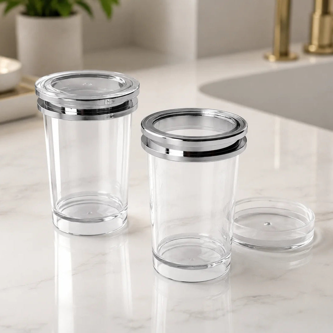Two clear glass storage jars with shiny silver metal rims and lids, shown on a white marble countertop near a sink.