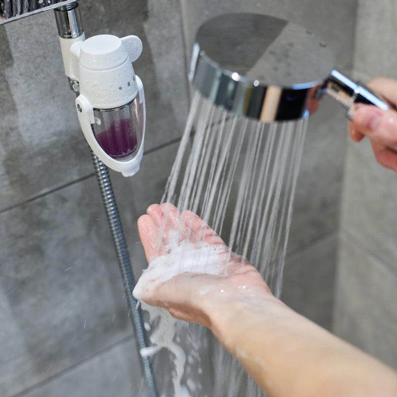 A Soap Shower soap dispenser mounted on a shower, dispensing white foam into a hand from a chrome shower head. The dispenser has a clear container with purple liquid soap inside and a white plastic body.